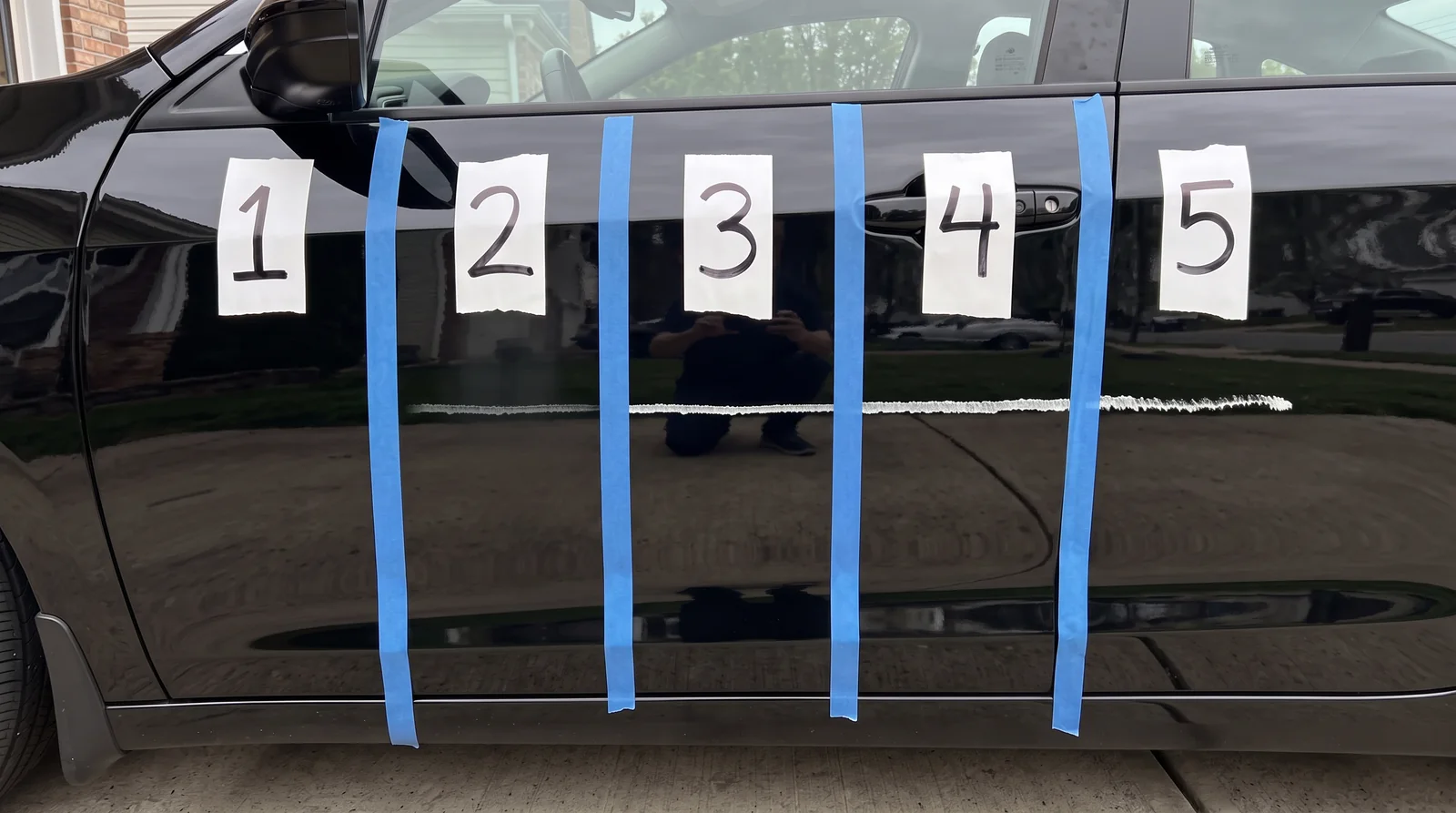 One key scratch across five taped test lanes on a black car door; only lane 1 is clean